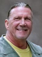 A man with slicked-back hair and a visible smile, wearing a green collared shirt over a yellow top, is posing in front of a grey background.